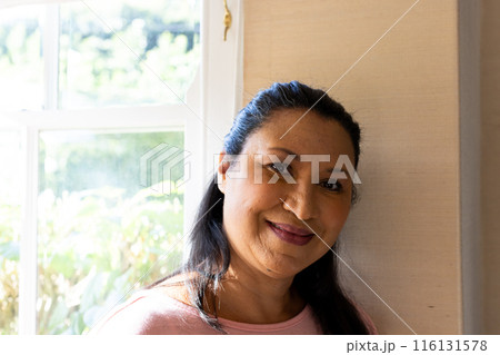 Mature biracial woman standing by window, looking calm at home 116131578