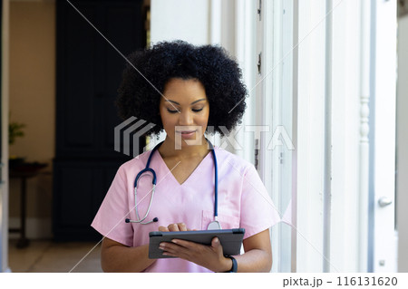 Young biracial female nurse wearing pink scrubs checking tablet at home 116131620