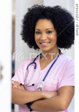 Biracial nurse standing with arms crossed, wearing scrubs at home 116131628