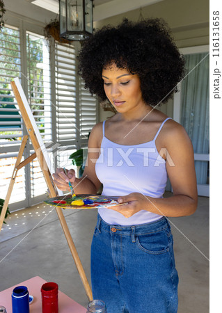 Biracial young woman painting on canvas at home, mixing colors on palette 116131658