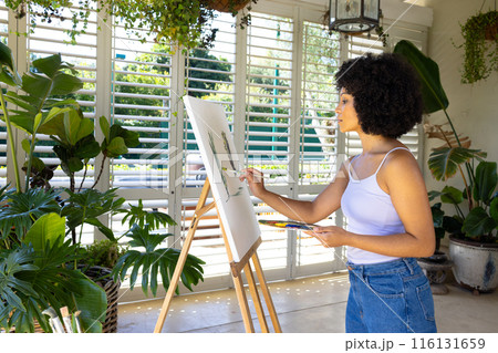 Young biracial woman painting on canvas in plant-filled room at home 116131659