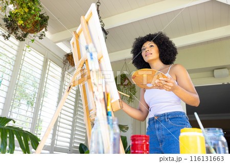 Young biracial woman painting on canvas at home, holding palette 116131663