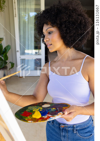 Young biracial woman holding palette, painting on canvas at home 116131667