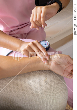 Mature biracial woman checking blood pressure at home, young biracial female nurse watching Mature biracial woman checking blood pressure at home, young biracial female nurse watching 116135004