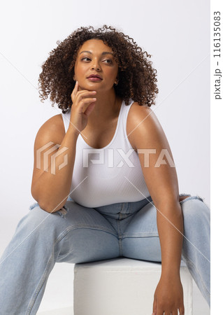 Biracial young female plus size model sitting, touching chin, looking thoughtful on white background 116135083