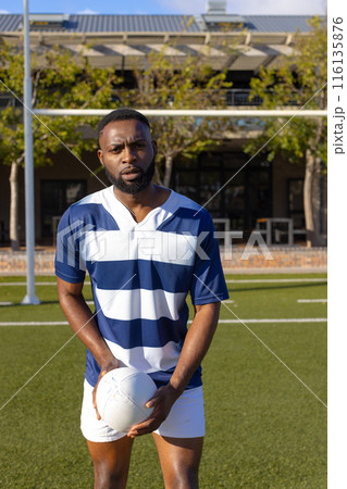 African American young male athlete holding rugby ball on field outdoors, looking serious African American young male athlete holding rugby ball on field outdoors, looking serious 116135876