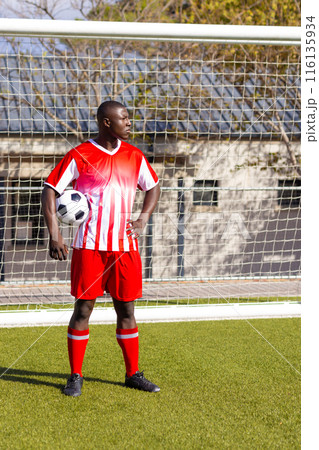 African American young male athlete holding a soccer ball, standing on field outdoors, copy space 116135934