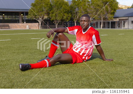 African American young male athlete sitting on grass on field outdoors, wearing red soccer uniform 116135941