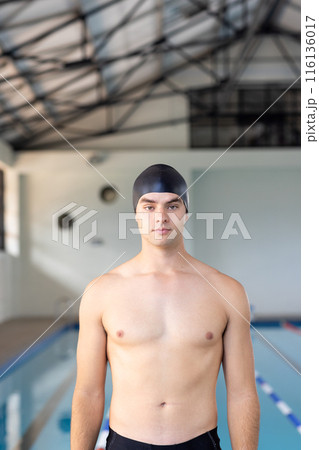 Caucasian young male swimmer standing by pool indoors, looking at camera, copy space 116136017