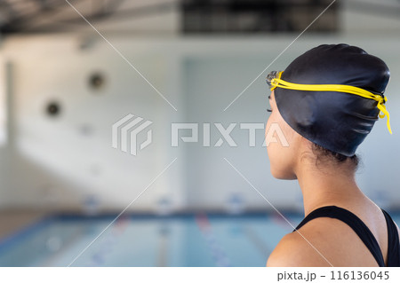 Biracial young female swimmer wearing a black cap is looking at a pool indoors, copy space 116136045