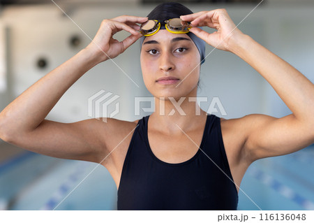 Biracial young female swimmer adjusting goggles indoors, ready to swim 116136048