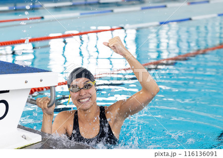 Biracial young female swimmer celebrating indoors in pool, wearing goggles 116136091