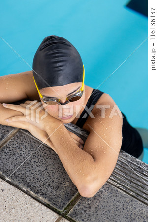 Biracial young female swimmer resting at pool edge indoors, wearing goggles Biracial young female swimmer resting at pool edge indoors, wearing goggles 116136137