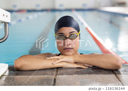 Young biracial female swimmer resting at pool edge indoors, wearing goggles 116136144