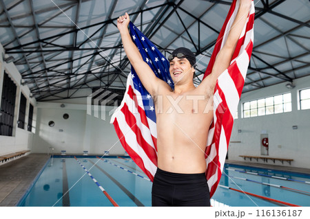 Caucasian young male swimmer holding American flag, standing by pool indoors, celebrating 116136187