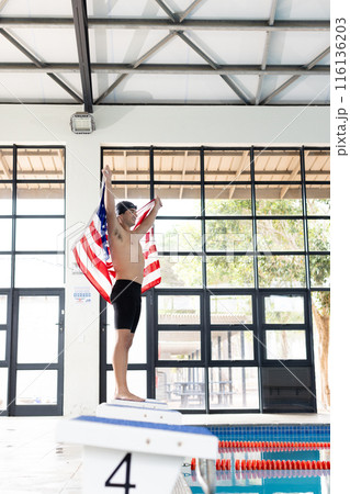 Caucasian young male swimmer holding American flag, standing on poolside indoors 116136203
