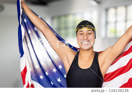 Biracial young female swimmer holding American flag indoors, wearing goggles, smiling Biracial young female swimmer holding American flag indoors, wearing goggles, smiling 116136236