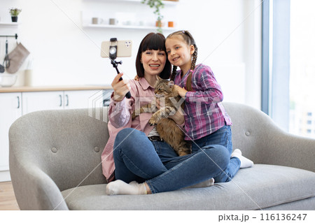 Mother and daughter taking selfie with cat on sofa 116136247