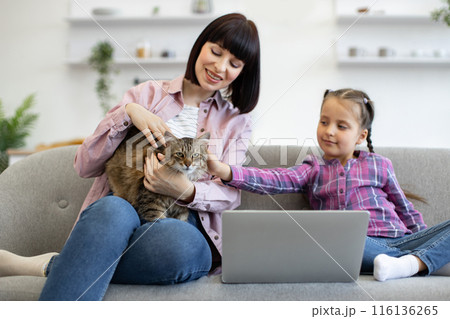 Mother and daughter with cat using laptop on cozy sofa 116136265