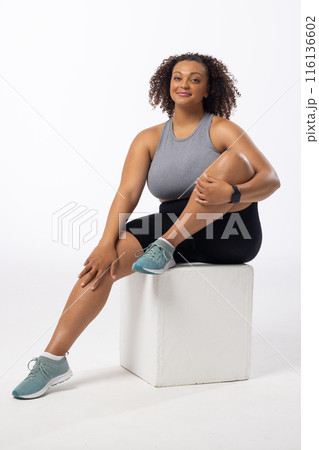 A biracial young female plus size model sits on white block, white background, copy space 116136602