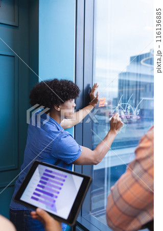 Young biracial man writing on graphs on window in a modern office Young biracial man writing on graphs on window in a modern office 116136885