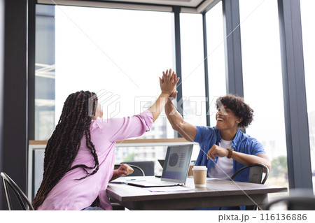 Diverse colleagues high-fiving at table with a laptop in a modern business office Diverse colleagues high-fiving at table with a laptop in a modern business office 116136886