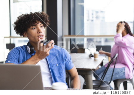 Biracial man with headphones talks on a smartphone, woman nearby in modern office 116136887