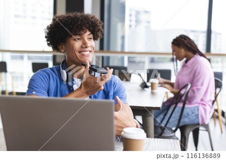 A young biracial man with curly hair is talking on a headset, working on a laptop in a modern busine 116136889