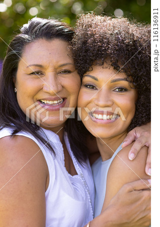 Mature biracial mother embraces her young daughter, both smiling in the garden Mature biracial mother embraces her young daughter, both smiling in the garden 116136911