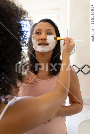 Mature biracial woman and daughter use facial masks, looking in mirror Mature biracial woman and daughter use facial masks, looking in mirror 116136917