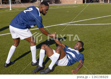Two African American male athletes play rugby, one helping the other up 116136920