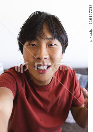 Asian teen boy with braces takes a selfie and video calls at home in a red shirt 116137252