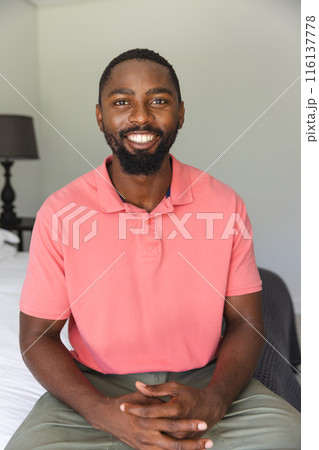 African American man sitting at home, having a video call, wearing a pink shirt, smiling 116137778