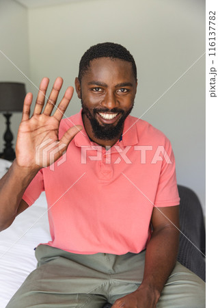 African American man waving during a video call, sitting on bed at home 116137782