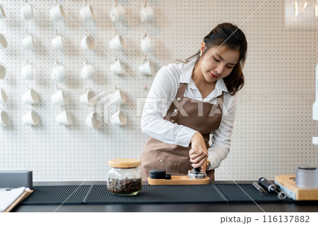An Asian female barista is making a coffee for customers in the coffee shop, pressing ground coffee. An Asian female barista is making a coffee for customers in the coffee shop, pressing ground coffee. 116137882