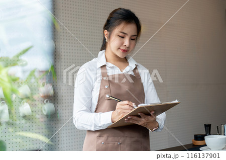 A young Asian female barista or waitress is working in a coffee shop, writing orders on the paper. A young Asian female barista or waitress is working in a coffee shop, writing orders on the paper. 116137901
