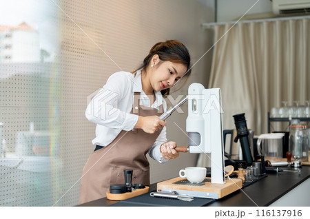 A beautiful young Asian female barista is working in a coffee shop, making a coffee with a machine. 116137916