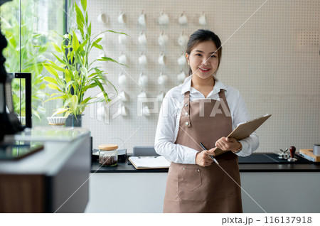 A charming Asian female barista in a brown apron is standing in a minimal white coffee shop. 116137918