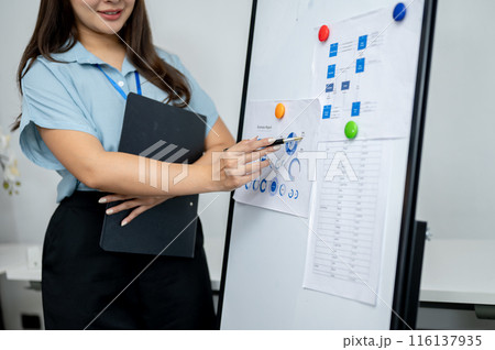 An Asian businesswoman is standing in the meeting, pointing her pen at a paper on the whiteboard. An Asian businesswoman is standing in the meeting, pointing her pen at a paper on the whiteboard. 116137935