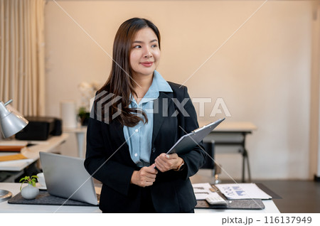 A charming Asian businesswoman holding a clipboard in her hand, is standing in the office. 116137949