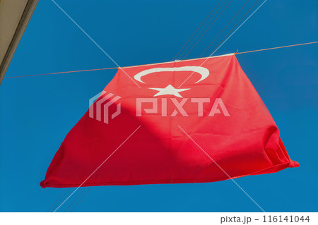 Turkey's red flag with a white crescent moon and a five-pointed star hangs on a rope between houses against a clear blue sky. Symbols of statehood, patriotism, signs, symbolism. Turkey's red flag with a white crescent moon and a five-pointed star hangs on a rope between houses against a clear blue sky. Symbols of statehood, patriotism, signs, symbolism. 116141044