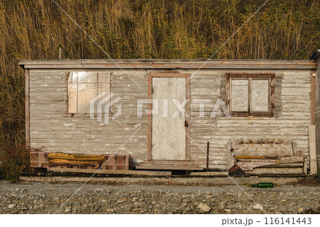 A weathered wooden shack with a central door, two windows, and an old couch outside. The background features tall, dry grass. 116141443