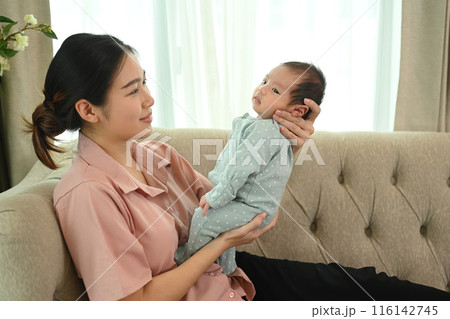 Shot of young Asian mother gently cradling her infant sitting on couch. Child care and motherhood concept 116142745
