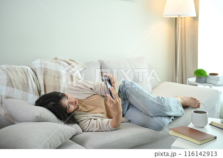 Pretty young Asian woman using mobile phone lying on couch in living room Pretty young Asian woman using mobile phone lying on couch in living room 116142913