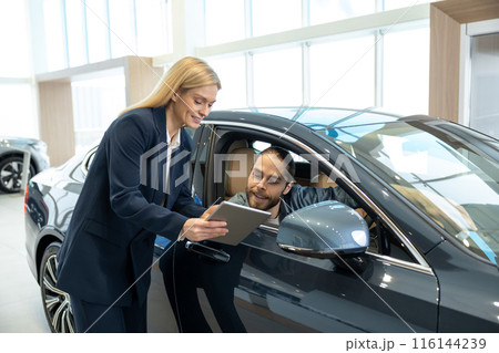 Blonde sale assistant showing something on a tablet to a customer in a dealership 116144239