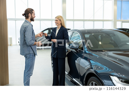 Female sale assistant in a dealership showing a new car to the customer Female sale assistant in a dealership showing a new car to the customer 116144329