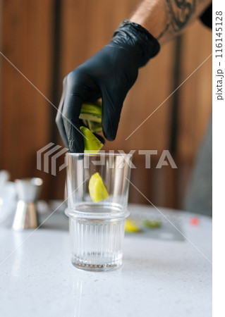 Closeup vertical cropped shot of unrecognizable bartender male in black gloves putting kiwi slices into glass to make deliciously refreshing cocktail in summer cafe. Concept of celebration mood. 116145128
