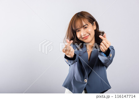 Confident business woman stands with a smile, creating a mini heart sign with her finger on a white background. Asian portrait of a beautiful young female sending love for Valentine's Day, studio shot 116147714