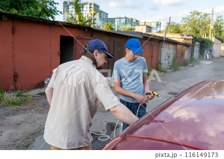 Dad and teenage son work together on the street near the garage. Outdoor activity. Family spend time. Helping and teach. Washing the car with a hand pump and hose. Bottled water. Dad and teenage son work together on the street near the garage. Outdoor activity. Family spend time. Helping and teach. Washing the car with a hand pump and hose. Bottled water. 116149173