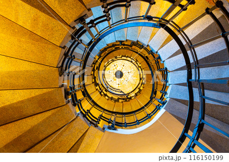 A view from the bottom of a winding staircase, looking up toward a chandelier. The staircase features a wooden handrail and metal railings. The surrounding walls are a warm, pale yellow color. A view from the bottom of a winding staircase, looking up toward a chandelier. The staircase features a wooden handrail and metal railings. The surrounding walls are a warm, pale yellow color. 116150199